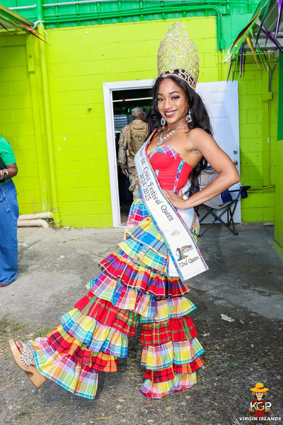 A festival queen in a red and madras gown poses in front of a green building.