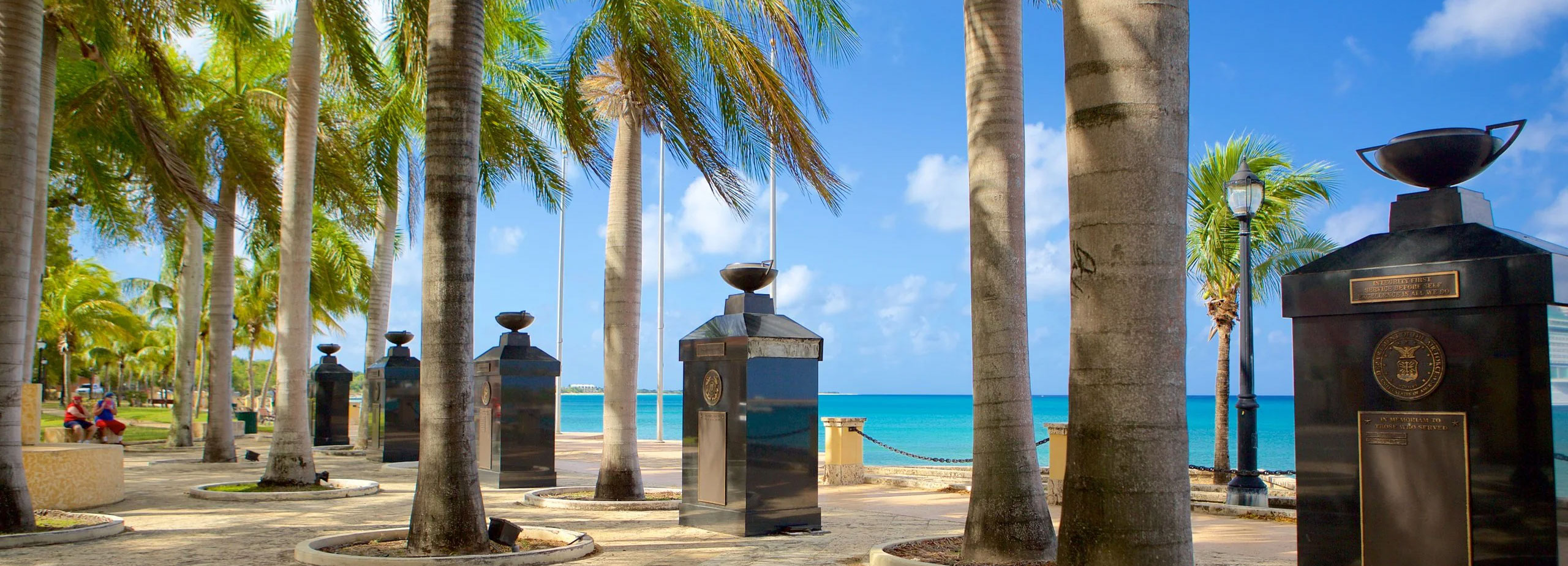 A sunlit, paved waterfront park in Frederiksted, featuring a row of tall palm trees interspersed with large, black pillar-style veterans memorials, with the bright turquoise Caribbean sea in the background.